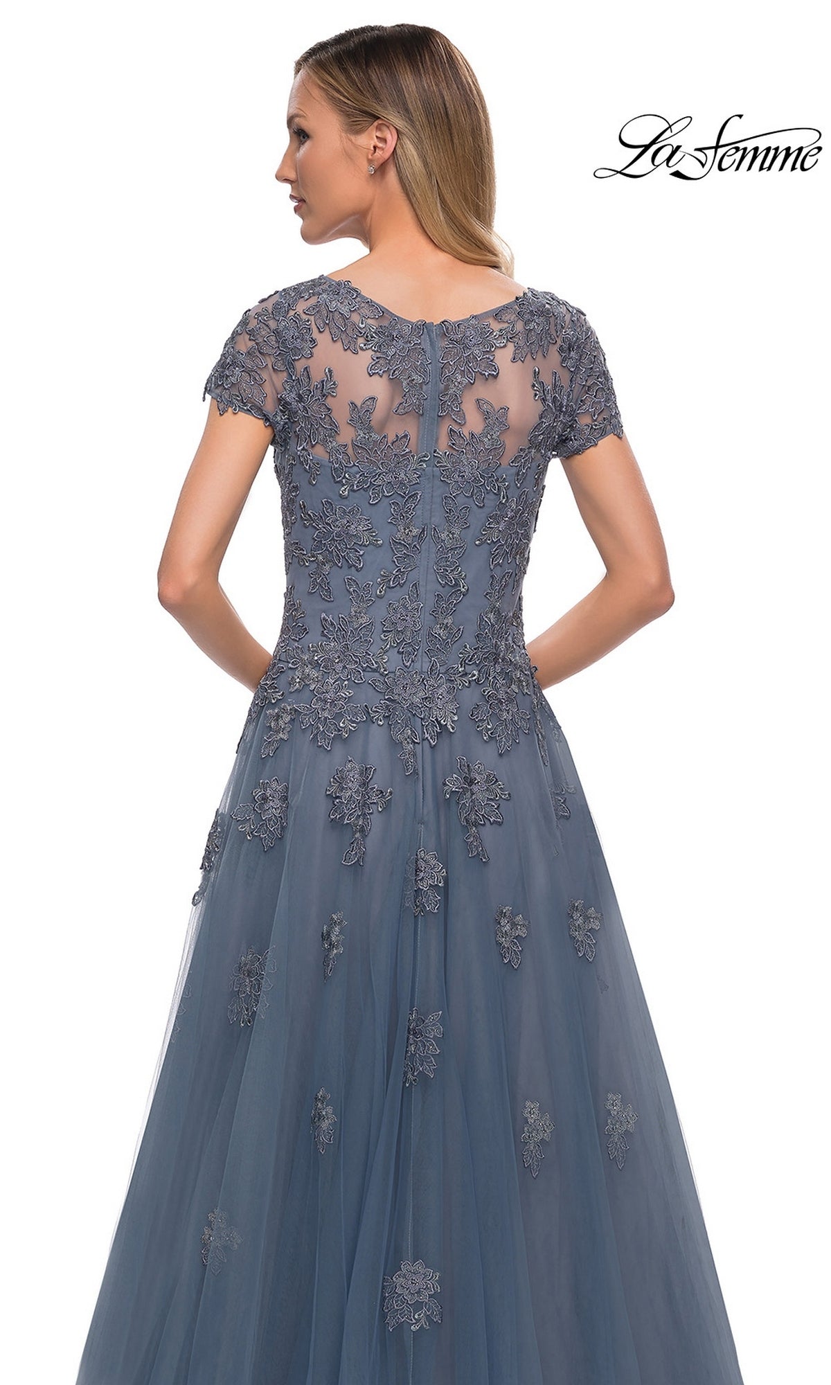 A woman in a blue Long Formal Dress: La Femme 29380 with lace appliques and floral embroidery stands facing away. The “La Femme” logo is in the top right corner.